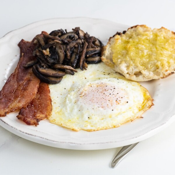 a fried egg over medium with bacon, sauteed mushrooms, and half an english muffin on a plate with a fork