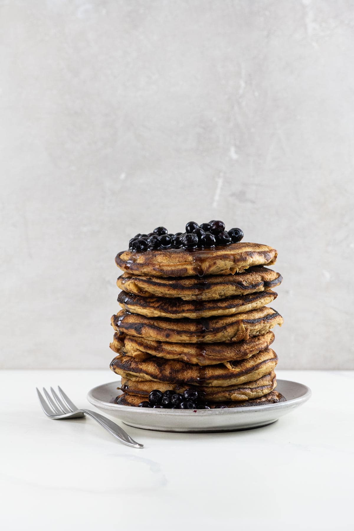 vegan banana pancakes stacked on a plate with blueberries, maple syrup, and a fork
