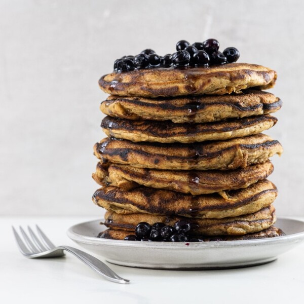 vegan banana pancakes stacked on a plate with blueberries, maple syrup, and a fork