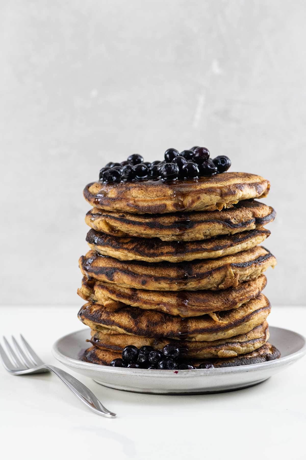 vegan banana pancakes stacked on a plate with blueberries, maple syrup, and a fork