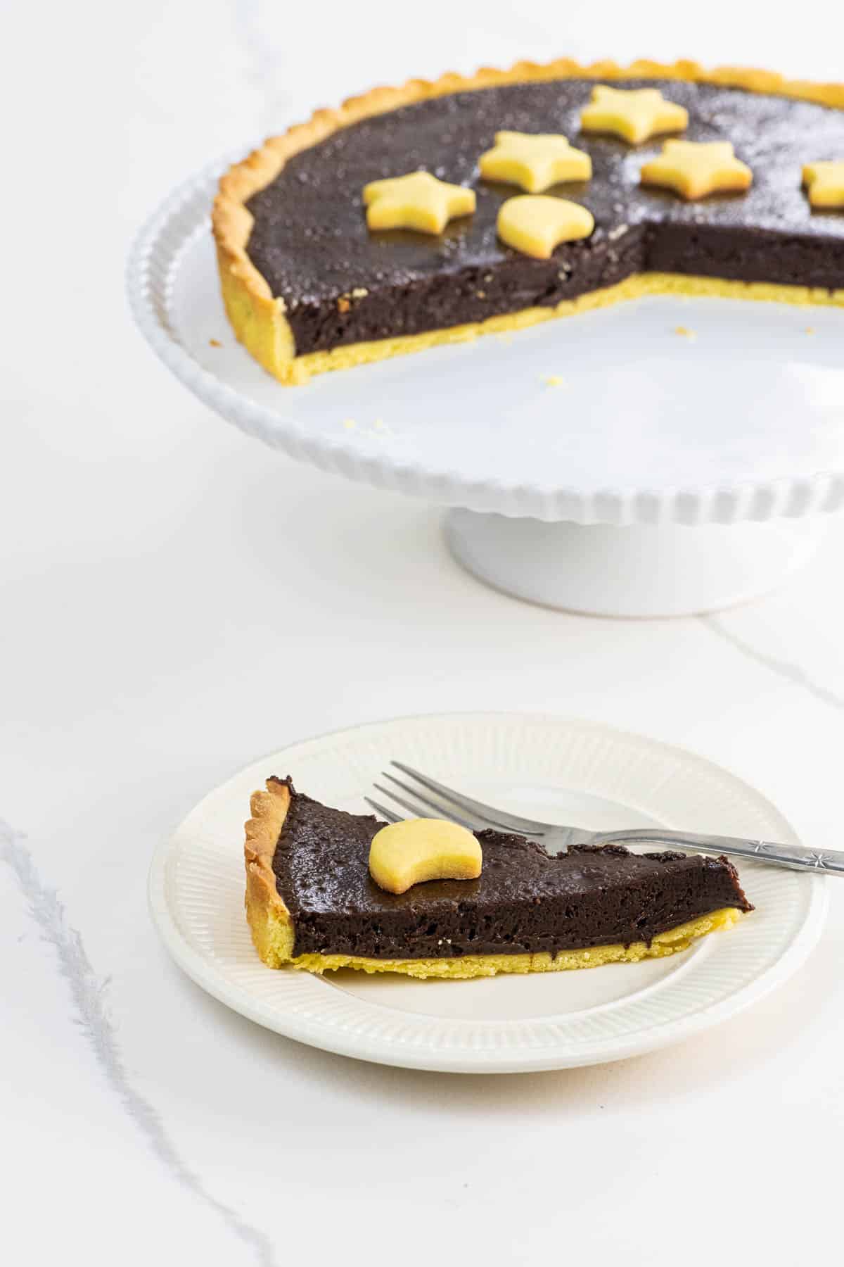 a bittersweet chocolate ganache tart with shortbread crust on a cake stand with a slice on a plate