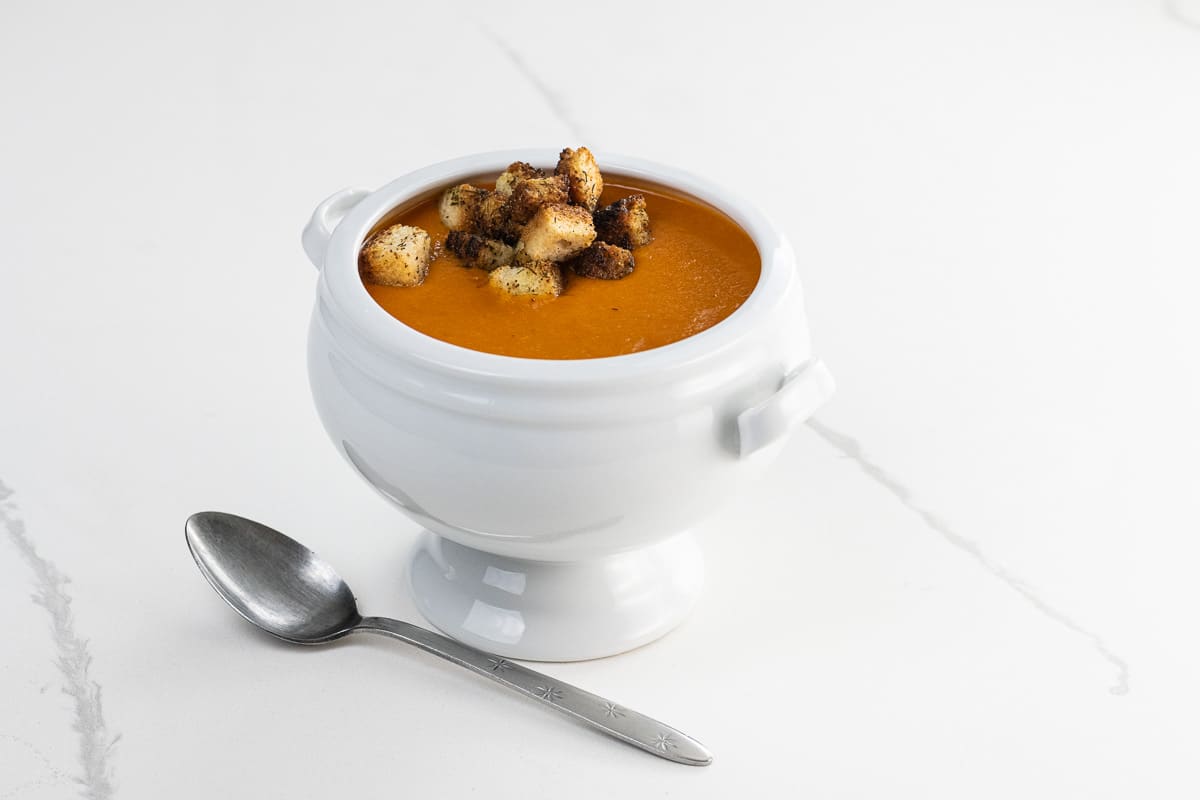 creamy vegan tomato soup with croutons in a footed bowl with a spoon