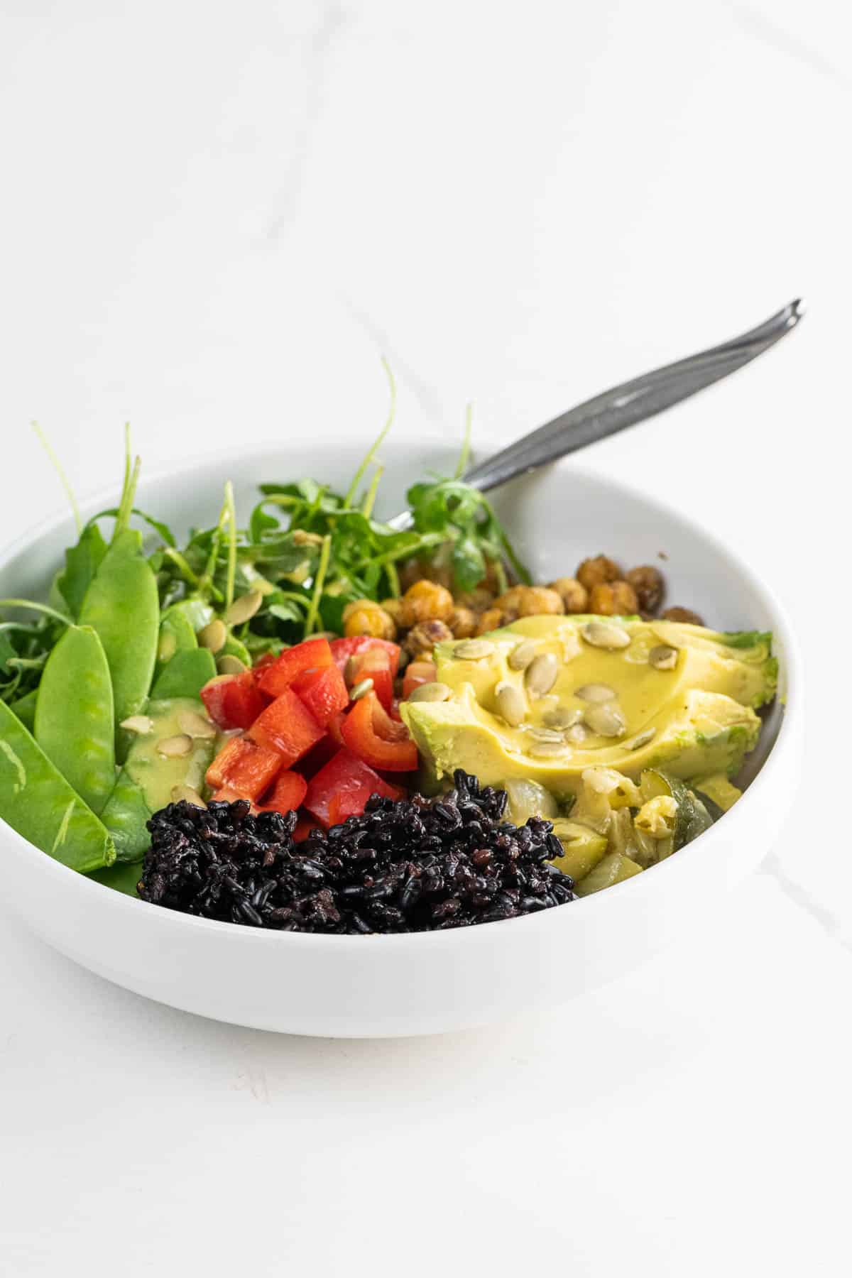 a vegan buddha bowl with forbidden rice, avocado, sauteed chickpeas, snow peas, red bell pepper, arugula, creamy lemon vinaigrette, and pepitas