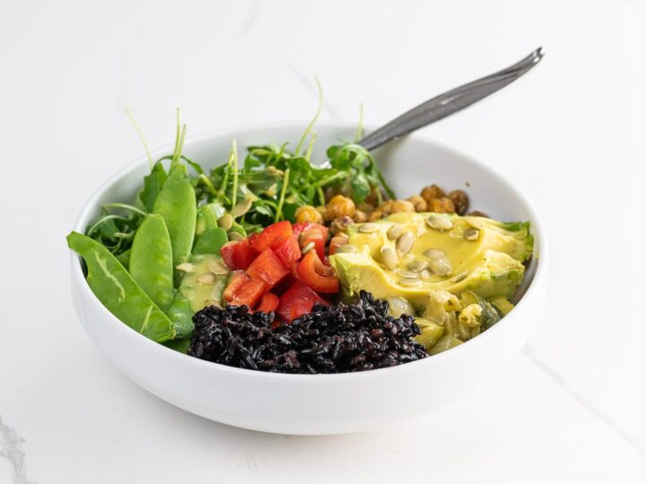 a vegan buddha bowl with forbidden rice, avocado, sauteed chickpeas, snow peas, red bell pepper, arugula, creamy lemon vinaigrette, and pepitas