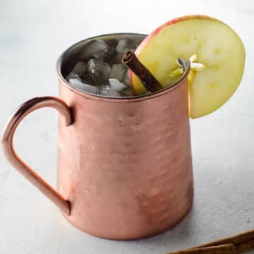 an apple cider moscow mule in a copper mug, garnished with an apple slice and a cinnamon stick