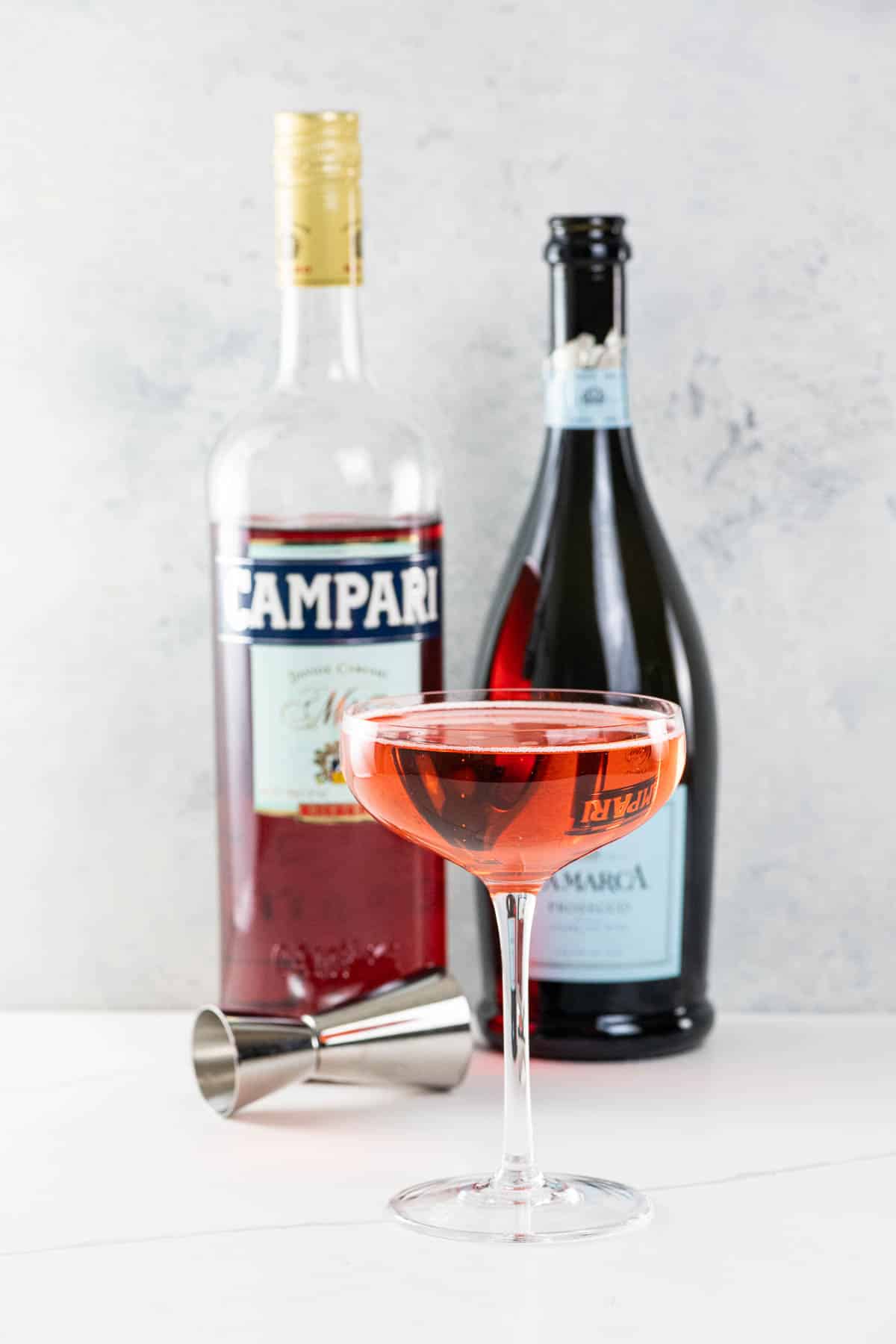 a campari prosecco cocktail (Mom's Italian soda) in a coupe glass with prosecco and campari bottles and a jigger in the background
