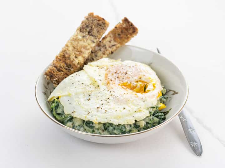 a fried egg over creamed spinach with toast soldiers in a shallow bowl with a fork