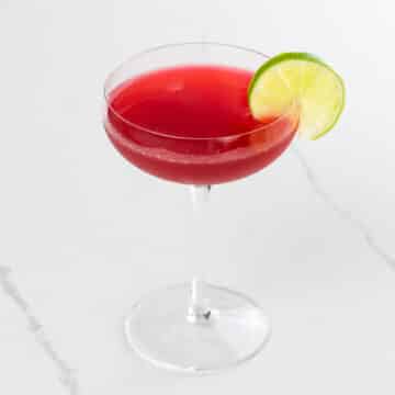 a raspberry gimlet in a coupe glass garnished with a lime wheel
