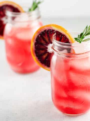 two blood orange cocktails in mason jars, each garnished with a rosemary sprig and a blood orange wheel
