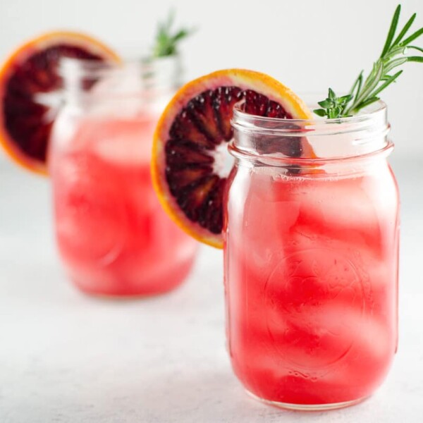 two blood orange cocktails in mason jars, each garnished with a rosemary sprig and a blood orange wheel