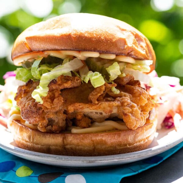 a crispy chicken sandwich on a plate on the arm of an adirondack chair with foliage in the background and a paper napkin
