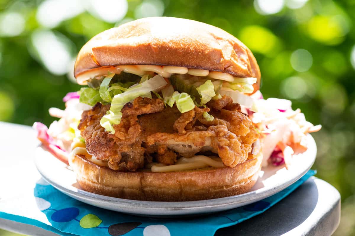 a crispy chicken sandwich on a plate on the arm of an adirondack chair with foliage in the background and a paper napkin