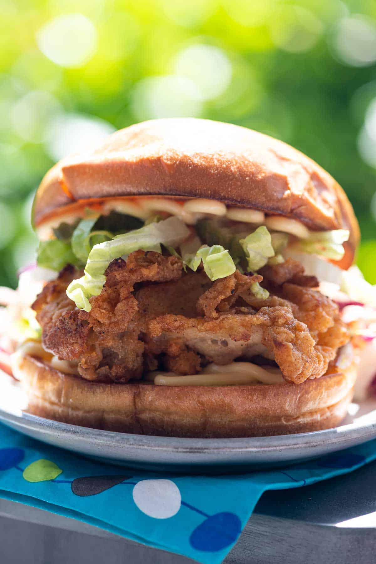 a crispy chicken sandwich on a plate on the arm of an adirondack chair with foliage in the background and a paper napkin