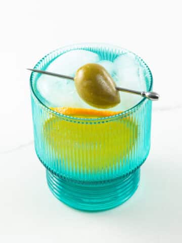 a blue glass with blanco vermouth and soda, a skewered olive, and an orange slice