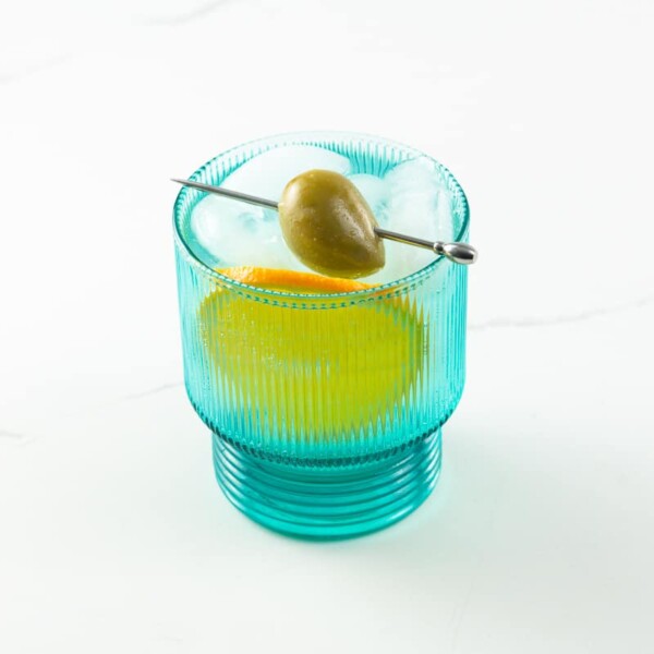 a blue glass with blanco vermouth and soda, a skewered olive, and an orange slice