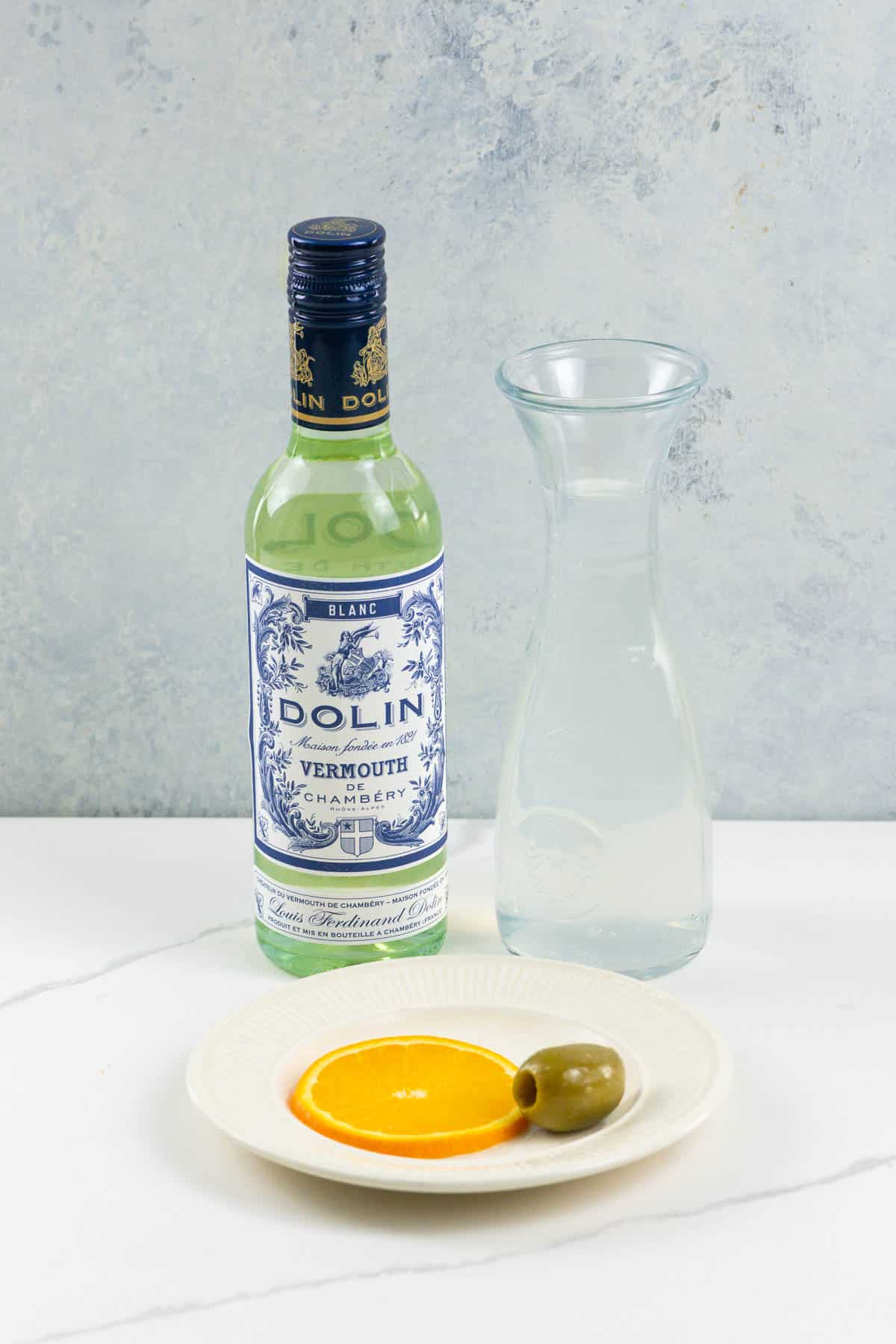 a bottle of dolin blanco vermouth, a carafe of soda water, an olive and an orange slice