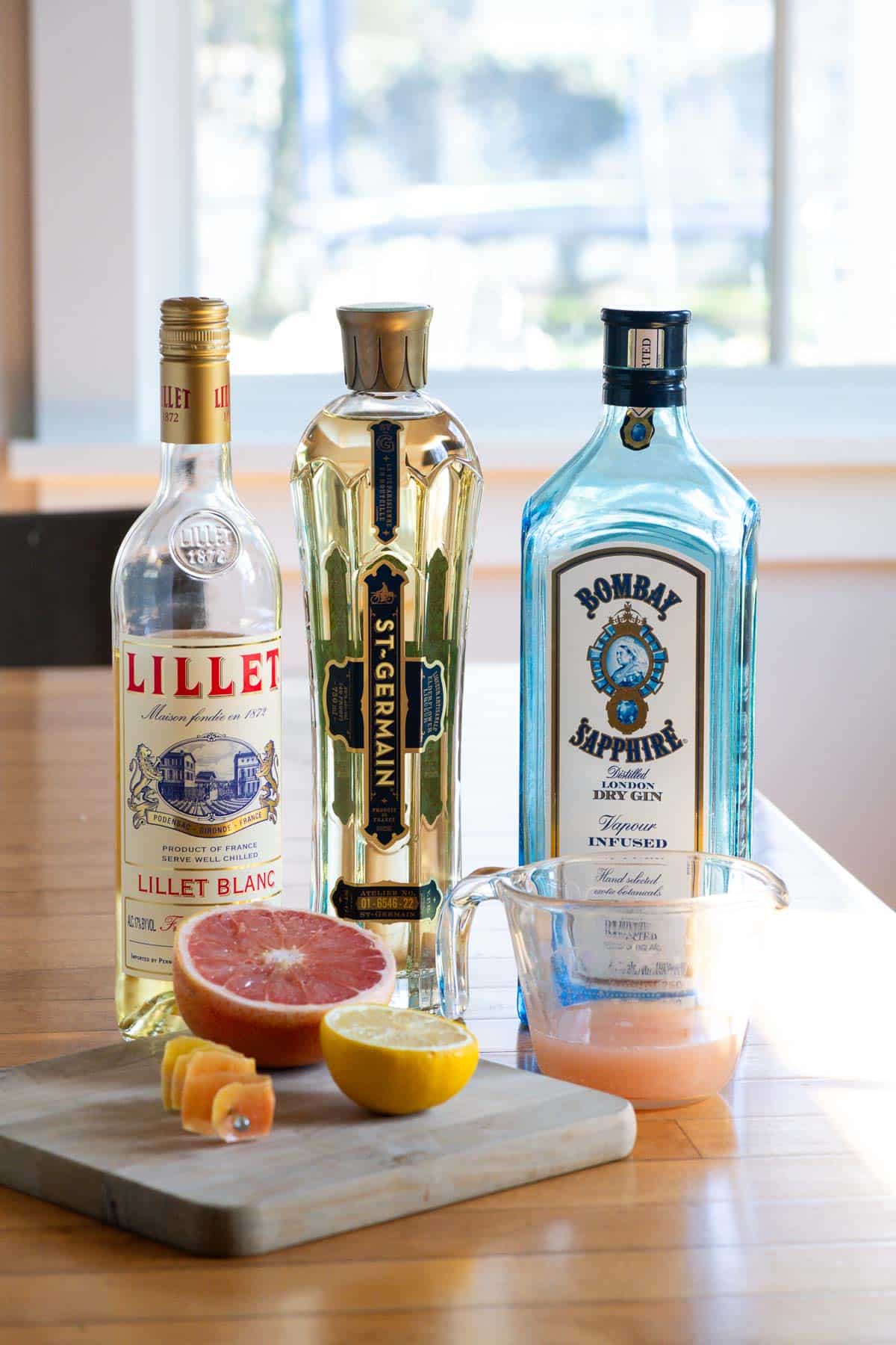 bottles of lilet blanc, st-germain, bombay sapphire gin, and grapefruit juice, a grapfruit half, and a lemon half